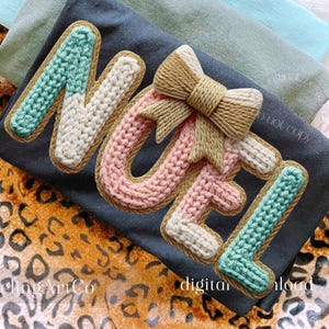 May include: A dark gray t-shirt with the word "NOEL" in a knitted design. The letters are in shades of blue, pink, and white, with a gold bow and trim. The words "Feeling ArtCo" and "digital download" are also visible.