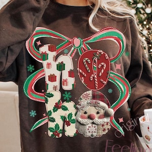 Può includere: Una felpa marrone con un design a tema natalizio. Il design include la scritta "HO HO HO" con scatole regalo, bastoncini di zucchero e una faccia di Babbo Natale. Un grande fiocco rosso e verde è dietro il testo. La felpa è a girocollo.