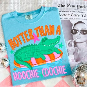 Op de afbeelding: Lichtblauw T-shirt met de tekst "HOTTER THAN A HOOCHIE COOCHIE" in oranje en roze. Het shirt heeft een groene alligator die op een roze en oranje zwemband drijft. Merk Current Colors.