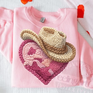 May include: A light pink sweatshirt featuring a heart design with a cowboy hat on top. The heart is a deep pink with a lighter pink design. The cowboy hat is tan and brown. Scattered around the sweatshirt are heart-shaped confetti.
