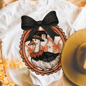 Peut inclure: T-shirt blanc avec un dessin d'oie de dessin anim&eacute; portant un chapeau de sorci&egrave;re et un col &agrave; volants, encadr&eacute; d'un n&oelig;ud noir. Un chapeau jaune et des fleurs sont &agrave; c&ocirc;t&eacute; du t-shirt. Le design est sur le th&egrave;me d'Halloween.