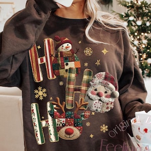 Op de afbeelding: Bruin sweatshirt met een feestelijk "HO HO HO" ontwerp. Het ontwerp bevat een sneeuwpop, de kerstman en een rendier, samen met sneeuwvlokken en sterren. De sweatshirt heeft een ronde hals en lange mouwen, perfect voor de feestdagen.