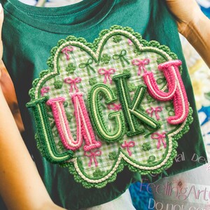 May include: A green t-shirt with the word "LUCKY" in an embroidered design. The design features a clover and bow pattern with pink and green accents. The shirt is held up by a person.