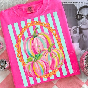 May include: A bright pink Comfort Colors sweatshirt featuring a colorful pumpkin design. The pumpkins are painted with pink, yellow, and blue stripes, set against a light blue striped background. The design is framed by an orange scalloped border.