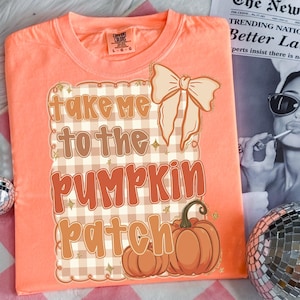 May include: Peach-colored t-shirt with the phrase "Take Me To The Pumpkin Patch" in a fall-themed design. The design includes a bow, pumpkins, and a plaid pattern. The shirt is a Comfort Colors brand.