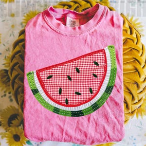 May include: A pink t-shirt with a watermelon slice applique. The watermelon design features a red and white checkered pattern, green rind, and black seeds. The shirt is folded on a yellow pillow with a floral background.