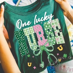 May include: Green t-shirt with the text "One lucky NURSE" in white and patterned letters. The design includes a stethoscope, shamrocks, and horseshoes. The shirt is held up by a person.