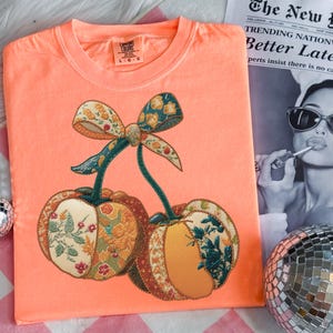May include: Coral-colored t-shirt featuring an embroidered design of two cherries with a bow. The cherries have a patchwork design with floral patterns in shades of orange, green, and cream. The shirt has a crew neck and short sleeves.