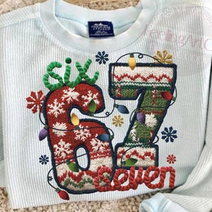 May include: Light blue long-sleeved shirt with a festive design. The design features the numbers "67" in a Christmas sweater style, with snowflakes, lights, and the word "seven" in red. The word "six" is in green.