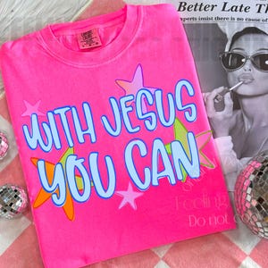 Könnte beinhalten: Ein leuchtend neonpinkes T-Shirt mit dem Schriftzug "WITH JESUS YOU CAN" in hellblauer Schrift mit dunkelblauem Rand. Das Design ist mit mehreren bunten Sternen in Rosa-, Orange- und Grüntönen verziert, die auf die Vorderseite gedruckt sind.
