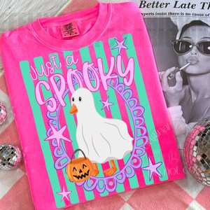 May include: A bright pink t-shirt featuring a graphic of a white ghost duck wearing a sheet, holding an orange jack-o'-lantern bucket. The design includes teal vertical stripes, purple stars, a purple oval frame, and the text Just a SPOOKY.