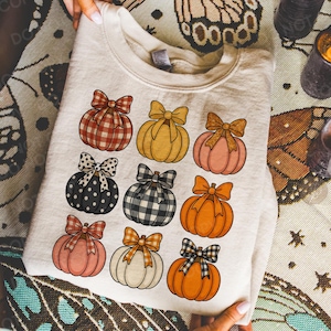 May include: A cream-colored sweatshirt featuring nine pumpkin illustrations with bows in various patterns and colors. The pumpkins are arranged in three rows of three. The background is a patterned fabric with candles.