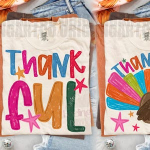 May include: Two cream-colored t-shirts featuring colorful graphic designs. The left shirt displays the word THANKFUL in multi-colored, distressed-style lettering with small stars. The right shirt shows the word THANKFUL above a brown turkey with a vibrant, multi-colored fan-shaped tail and small stars.