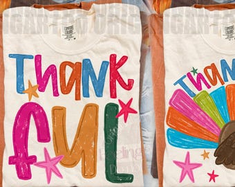 May include: Two cream-colored t-shirts featuring colorful graphic designs. The left shirt displays the word THANKFUL in multi-colored, distressed-style lettering with small stars. The right shirt shows the word THANKFUL above a brown turkey with a vibrant, multi-colored fan-shaped tail and small stars.