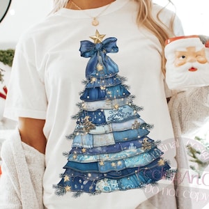 Könnte beinhalten: Weißes T-Shirt mit einem blauen Weihnachtsbaum-Design. Der Baum besteht aus geschichtetem, jeansähnlichem Stoff, verziert mit einer blauen Schleife, goldenen Sternen und einem Sternaufsatz. Ein festliches Urlaubsdesign.
