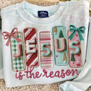 May include: Light blue sweatshirt with the words "JESUS is the reason" embroidered in pink. The letters are on decorative panels with various patterns and colors, including green plaid, red stripes, and pink with Christmas trees. A pink bow and a teal bow are on the panels.