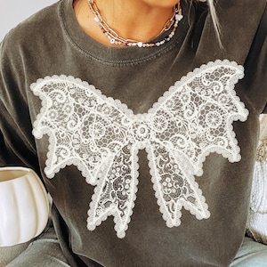 May include: Dark gray t-shirt featuring a large white lace bow design. The shirt has short sleeves and a crew neck. The model is wearing light blue jeans and holding a white pumpkin-shaped mug. The shirt is a casual, comfortable style.