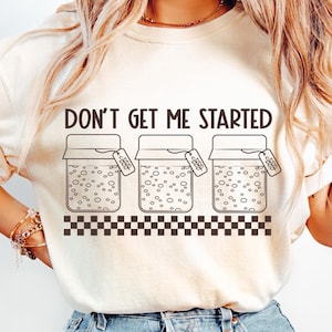 May include: A white t-shirt with the text "Don't get me started" and a graphic of three jars with lids and labels that say "good vibes". The jars are filled with bubbles and are sitting on a black and white checkered pattern.