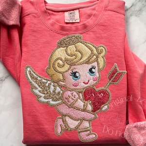 May include: Coral sweatshirt featuring an embroidered Cupid design holding a heart and arrow. The Cupid has blonde hair, blue eyes, and gold detailing. The cuffs are a lighter pink. The brand tag reads "Comfort Colors".