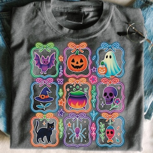 May include: Gray t-shirt featuring Halloween-themed graphics. The shirt displays nine colorful illustrations of a bat, pumpkin, ghost, witch hat, cauldron, skull, black cat, spider, and fish, each within a neon-outlined frame. The shirt is paired with blue jeans.