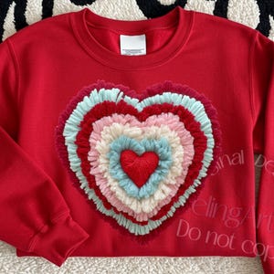 May include: A red sweatshirt with a multi-layered heart design in shades of pink, blue, and white. The sleeves have embroidered text. The sweatshirt is on a white rug with black flame-like shapes.