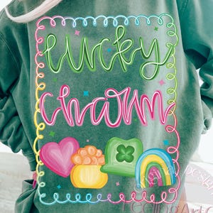 May include: Green sweatshirt with the words "Lucky Charm" in a colorful, whimsical design. The design includes a pink heart, a pot of gold, a green shamrock, and a rainbow. The sweatshirt is a comfortable, casual style.