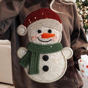 May include: A brown sweatshirt featuring a sequined snowman design. The snowman has a red hat, green scarf, and a carrot nose. The design is embroidered with sequins and black buttons. A festive winter holiday theme.