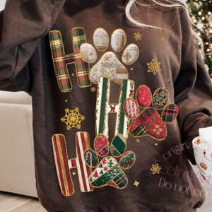 May include: A brown sweatshirt with the word "Ho Ho Ho" in a plaid and patterned design. The design includes paw prints and snowflakes. The sweatshirt is a festive holiday item.