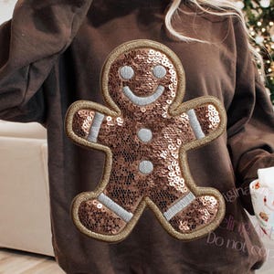 May include: A brown sweatshirt with a sequined gingerbread man design. The gingerbread man has a smiling face and silver accents. The sweatshirt is a long-sleeved, crew neck style. A festive holiday apparel item.