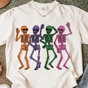 Puede incluir: Camiseta de color crema con un dise&ntilde;o de cuatro esqueletos bailando en naranja, morado, verde y rosa. Cada esqueleto lleva gafas de sol. La camiseta tiene cuello en V y mangas cortas. Se ve un collar plateado.