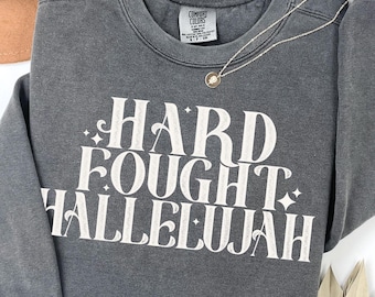 May include: A gray Comfort Colors sweatshirt with the words "HARD FOUGHT HALLELUJAH" in white script. A gold necklace and a brown hat are also visible. The sweatshirt has long sleeves and a crew neck.