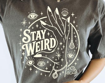 May include: Dark gray t-shirt with the words "STAY WEIRD" in a circular design. The design includes a hand, eyes, stars, and a pendant. The shirt is a short-sleeved, casual style.