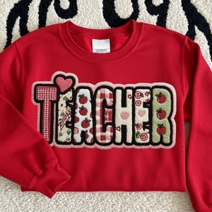 May include: A red cropped sweatshirt with the word "TEACHER" appliqued across the front. The letters are a mix of patterns, including gingham, hearts, and apples. The sweatshirt has long sleeves and a crew neck.