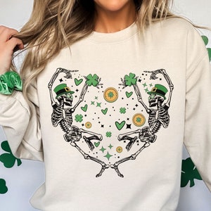 Op de afbeelding: Beige sweatshirt met lange mouwen met een hartvormig ontwerp van twee dansende skeletten met groene hoeden. Het ontwerp bevat groene klavers, gouden munten en groene sterren. Een casual stijl.