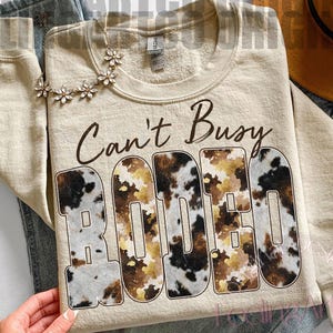Puede incluir: Sudadera beige con el texto "Can't Busy Rodeo" en escritura marrón. La palabra "RODEO" está en letras grandes con un estampado de piel de vaca. Se ven un collar floral y un sombrero.