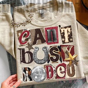 May include: A beige sweatshirt with the text "Can't Busy Rodeo" in a collage style. The design includes a horseshoe, a disco ball, and star accents. The sweatshirt is paired with blue jeans and a necklace.