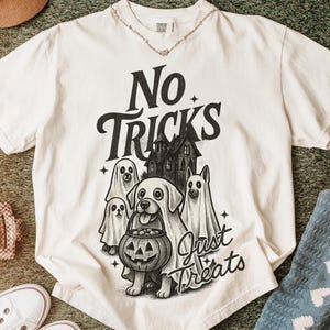 Op de afbeelding: Crèmekleurig T-shirt met een Halloween-thema. Het ontwerp toont een hond met een pompoen, geesten en een spookhuis. De tekst op het shirt luidt "No Tricks Just Treats".