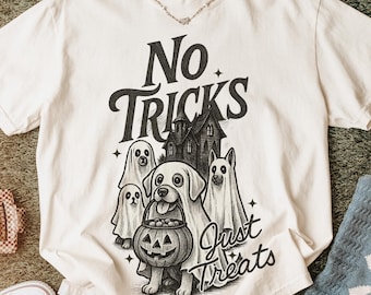 May include: Cream-colored t-shirt with a Halloween-themed graphic. The design features a dog holding a jack-o'-lantern, surrounded by ghosts and a haunted house. Text reads "No Tricks Just Treats".