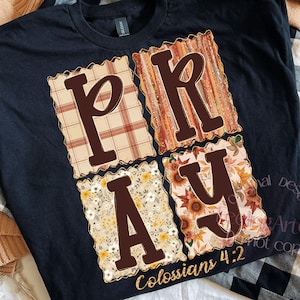 May include: Black t-shirt with the word "PRAY" in brown block letters, each letter within a decorative square. The squares feature plaid, floral, and sunflower patterns. The text "Colossians 4:2" is in gold script.
