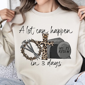 May include: A beige sweatshirt with the text "A lot can happen in 3 days" and a graphic of a cross with a crown of thorns, nails, and a stone with the text "He is Risen".