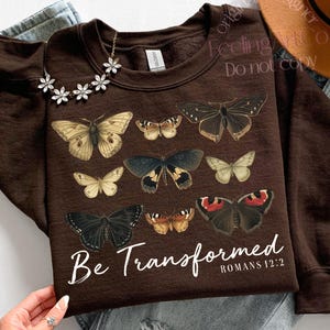 以下が含まれることがあります： 様々な蝶のイラストが描かれた茶色のスウェットシャツ。「Be Transformed」の文字と「Romans 12:2」の参照が白で印刷されています。花のネックレスと帽子が見えます。