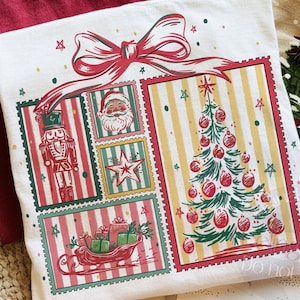 May include: White t-shirt with a Christmas-themed design featuring a large red bow and postage stamp-style illustrations of a nutcracker, Santa Claus, a Christmas tree, and a sleigh with gifts. The shirt is paired with a red shirt and a beige blanket.