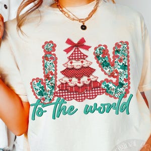 May include: Cream-colored t-shirt with a Christmas tree design. The tree is red and white plaid with a red bow at the top. The word "JOY" is in a floral pattern, and the phrase "To the world" is in teal script.