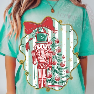 May include: A light teal t-shirt featuring a Christmas-themed graphic. The graphic includes a red nutcracker soldier, a Christmas tree, and a red bow. The design is framed with a gold glitter border and set against a striped background.