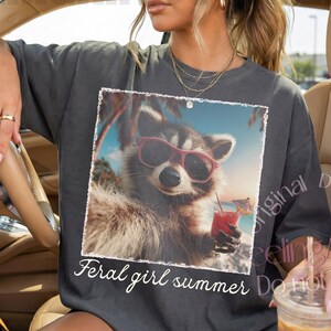 May include: Dark gray t-shirt featuring a raccoon wearing sunglasses and holding a tropical drink, with the text "Feral girl summer." The raccoon image is framed with a white border. The shirt is worn by a person in a car.