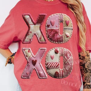 Könnte beinhalten: Korallfarbenes T-Shirt mit aufgenähten Buchstaben "XO" auf der Vorderseite. Die Buchstaben bestehen aus verschiedenen Stoffflicken in Rosa-, Rot- und Brauntönen mit Herzakzenten. Das Shirt hat eine lockere Passform und kurze Ärmel.