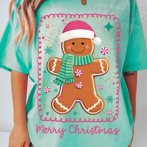 May include: A turquoise t-shirt with a gingerbread man design. The gingerbread man wears a pink Santa hat and green scarf, surrounded by candy and snowflakes. The words "Merry Christmas" are written below the design.
