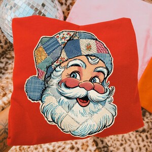 Op de afbeelding: Een rode trui met een afbeelding van een kerstman met een witte baard, blauwe ogen en een patchwork hoed. De kerstman staat centraal op de voorkant van de trui. De achtergrond bevat een discobal en kleurrijke kussens.