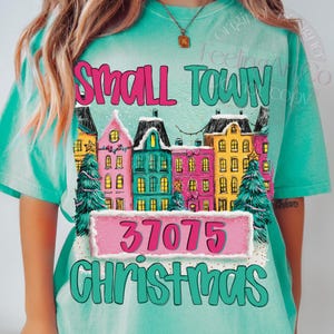May include: A turquoise t-shirt with a colorful graphic featuring the words "Small Town Christmas" and the number "37075". The graphic includes illustrations of buildings and Christmas trees.