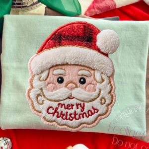 May include: A light green t-shirt with an embroidered Santa Claus face. The Santa has a red plaid hat, white beard, and the words "merry Christmas" embroidered in red. The shirt is folded on a red surface.